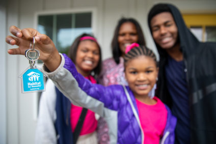 A Habitat for Humanity family in front of their new home; a smiling girl is holding a key with a Habitat keychain