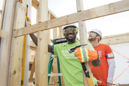 A volunteer is holding a hammer at a Habitat for Humanity home build site; in the background is a construction staff giving support