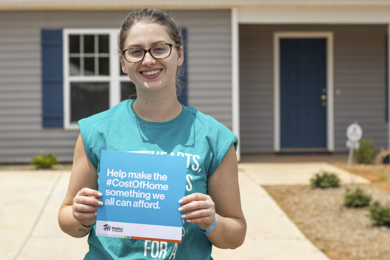 A smiling woman holding an advocacy statement card in front of a Habitat for Humanity house, "Helping make the Cost of Home something we can afford."